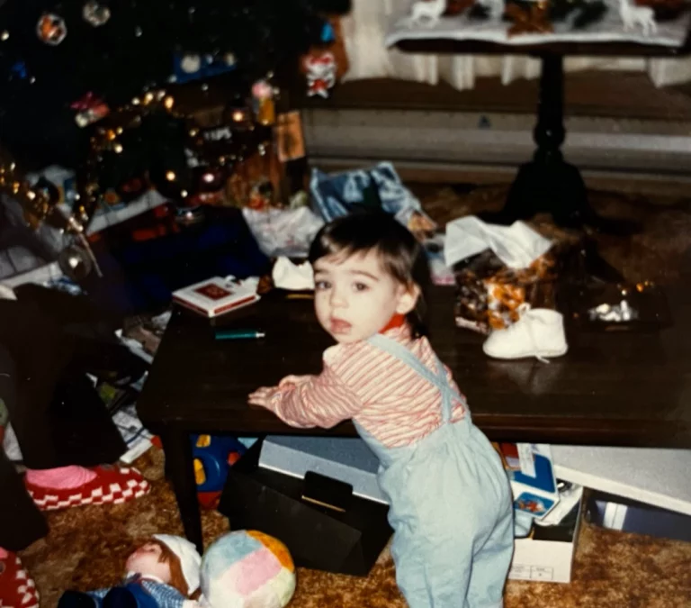 A photo of a living room and a toddler from the early 1980’s. The toddler looks to camera while holding on to a coffee table for balance. They wear overalls and striped shirt. The coffee table and the surrounding floor of the orange carpet are covered in toys and random objects.